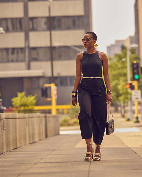 Fashion influencer Farotelle walking on a street while wearing an all-black outfit with gold heeled sandals and gold accessories.