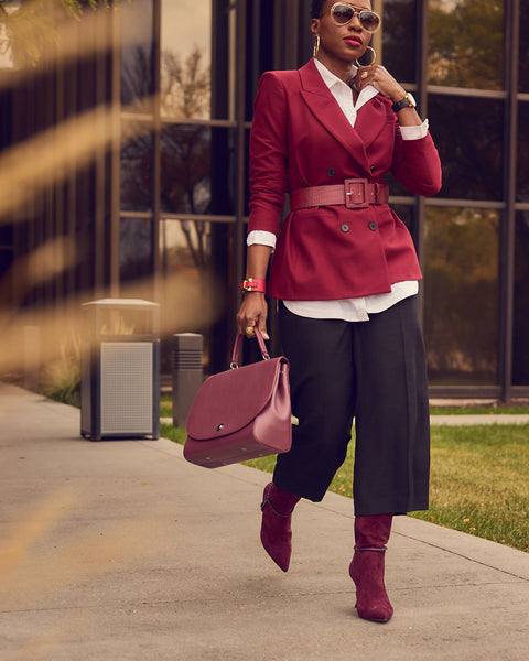 A fashion and style editorial photo showing influencer Farotelle in a business professional outfit. She is wearing black culottes with a white shirt under a belted burgundy blazer, and burgundy boots. She is also holding a classic burgundy leather satchel bag.