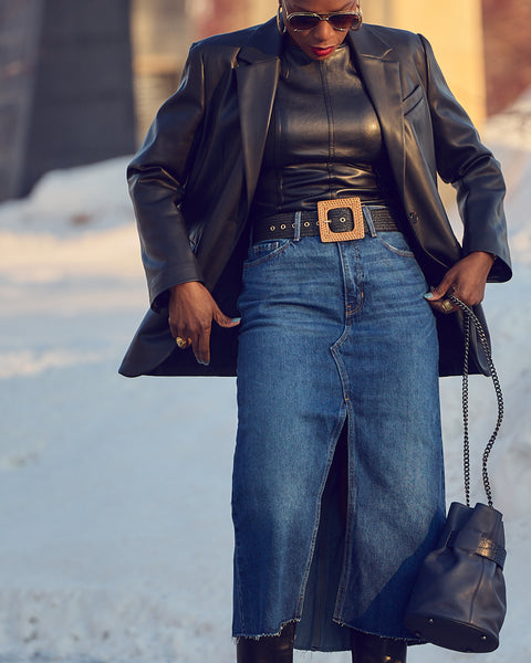 A woman wearing a blue denim skirt with a black faux leather blazer. She is holding a blue handbag with a chain strap.