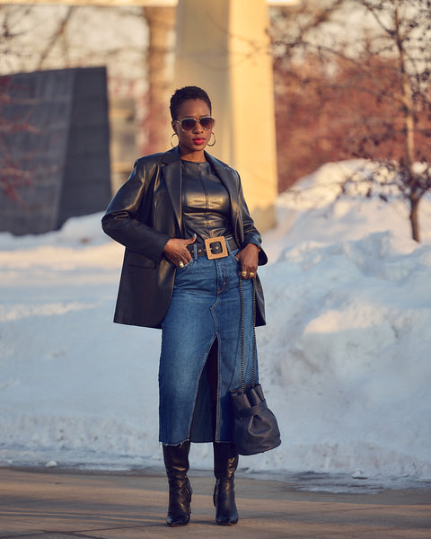 A woman wearing a blue denim skirt with a black faux leather blazer. She is also wearing black knee-high boots and holding a blue handbag.