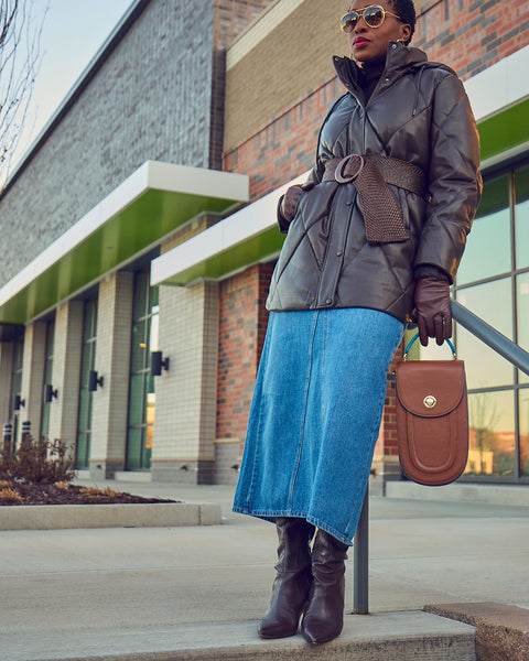 Style blogger Farotelle wearing a maxi blue denim skirt with a belted faux leather dark brown puffer coat. She is wearing dark brown boots that match her coat, and is holding a brown leather handbag.