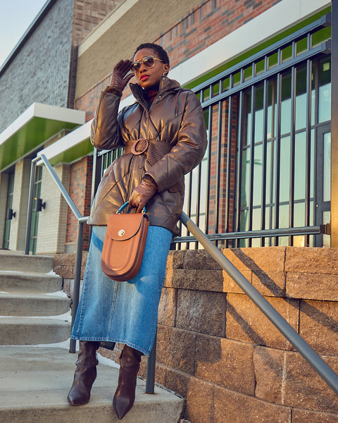Style blogger Farotelle wearing a maxi blue denim skirt with a belted faux leather dark brown puffer coat. She is wearing dark brown boots and leather gloves that match her coat, and is holding a brown leather handbag. She has on sunglasses.