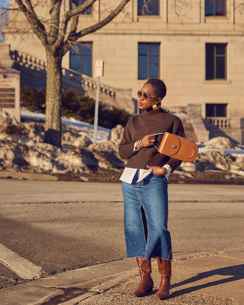 Style influencer Farotelle wearing a blue denim skirt and dark brown sweater layered over a white shirt. She has on tall brown boots and is holding a brown leather handbag. The outfit has a color-blocked effect.