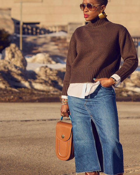 Style influencer Farotelle wearing a blue denim skirt and dark brown sweater layered over a white shirt. She is holding a brown leather handbag. The outfit has a color-blocked effect.
