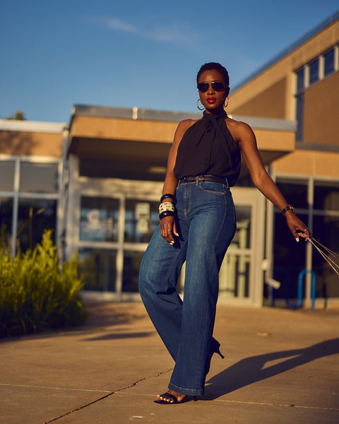 A fashion and style editorial photo showing influencer Farotelle wearing blue wide-leg jeans with a black halter top and black strappy sandals.