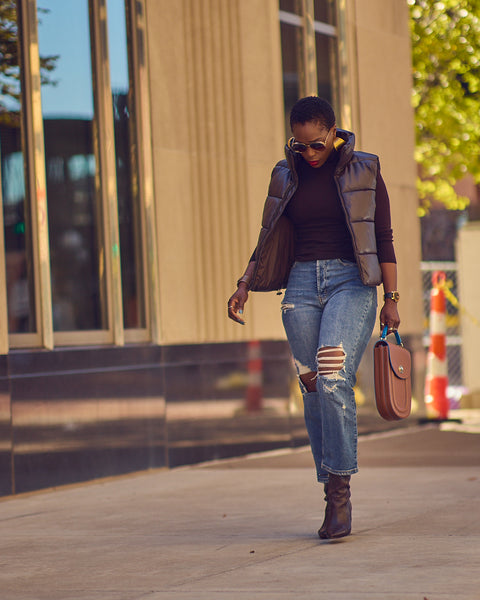 Style blogger Farotelle wearing distressed blue jeans with a dark brown turtleneck sweater and a dark brown faux leather puffer vest. She is also wearing dark brown boots and is holding a brown leather handbag.