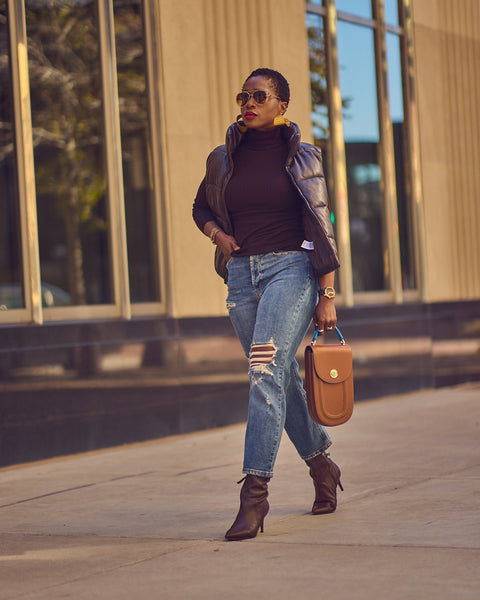 Style blogger Farotelle wearing distressed blue jeans with a dark brown turtleneck sweater topped with a dark brown faux leather puffer vest. She is also wearing dark brown boots and is holding a brown leather handbag.