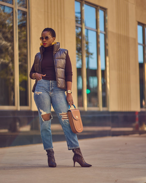 Style blogger Farotelle wearing blue mom jeans with a dark brown turtleneck sweater and a dark brown faux leather puffer vest. She is also wearing dark brown boots and is holding a brown leather handbag.