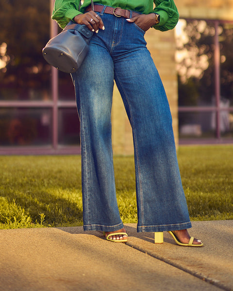 A fashion and style editorial photo showing influencer Farotelle wearing blue wide-leg jeans with a green blouse and green strappy sandals.