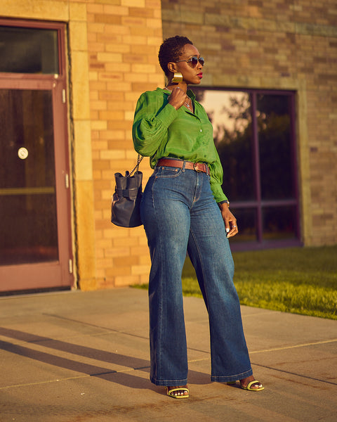 A fashion and style editorial photo showing influencer Farotelle wearing blue wide-leg jeans with a green blouse and green strappy sandals.