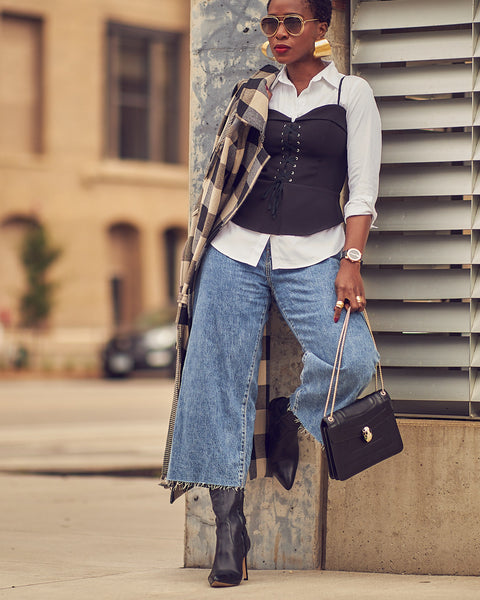 A woman wearing cropped blue jeans with a black corset top over a white shirt. She has on black boots and is holding a black handbag. She is wearing chunky gold earrings and sunglasses.