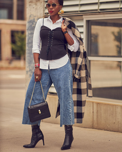 A woman wearing cropped blue jeans with a black corset top over a white shirt. She has on black boots and is holding a black handbag and a plaid coat. She is wearing chunky gold earrings and sunglasses.