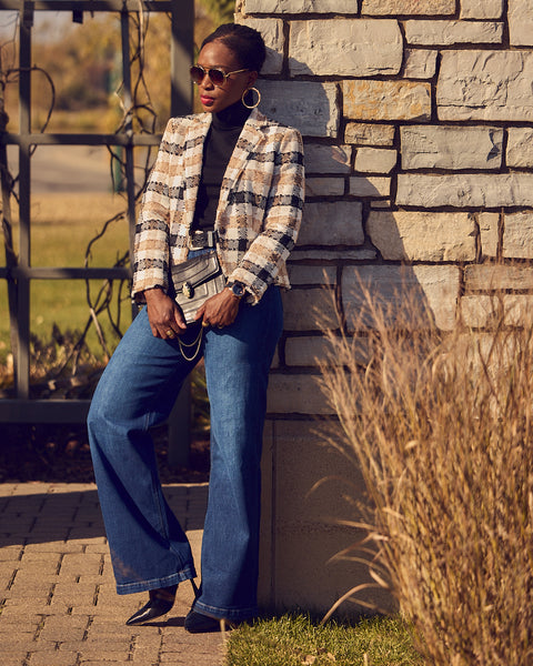 A woman wearing blue jeans with a black turtleneck and plaid cropped blazer. She is wearing black heels and is leaning against a brick wall.