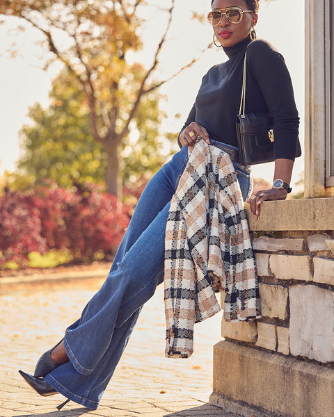 A woman wearing blue jeans with a black turtleneck. She is holding a plaid jacket and wearing black heels. She is leaning against a brick wall.