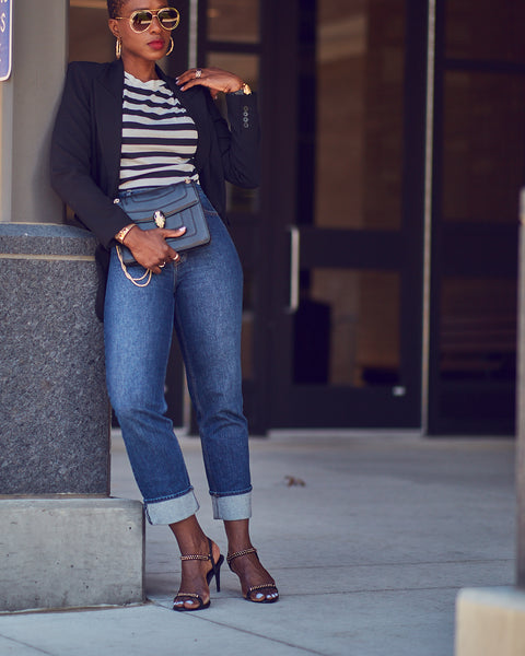 Fashion and style blogger Farotelle wearing an outfit that consists of high-waisted blue jeans, a striped tee and a black blazer. She is also wearing black strappy sandals and holding a black handbag. She has dark skin, short hair and is in a standing position.