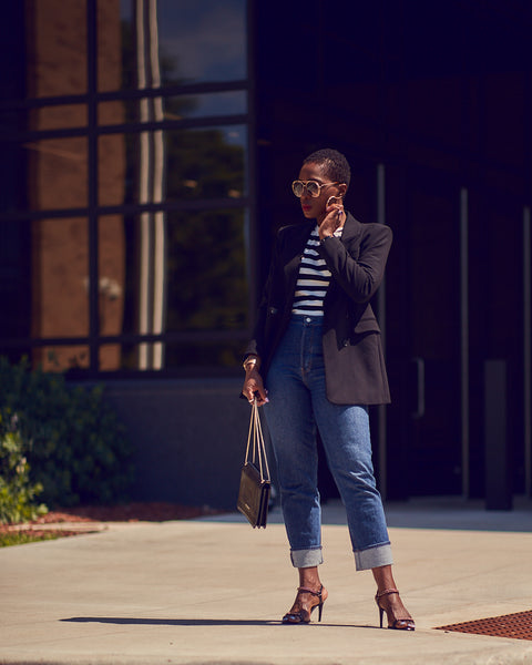 Fashion and style blogger Farotelle wearing an outfit that consists of high-waisted blue jeans, a striped tee and a black blazer. She is also wearing black strappy sandals and holding a black handbag. She has dark skin, short hair and is in a standing position.