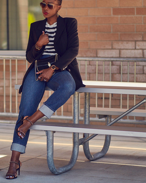 Fashion and style blogger Farotelle wearing an outfit that consists of high-waisted blue jeans, a striped tee and a black blazer. She is also wearing black strappy sandals and holding a black handbag. She has dark skin, short hair and is leaning against a table.