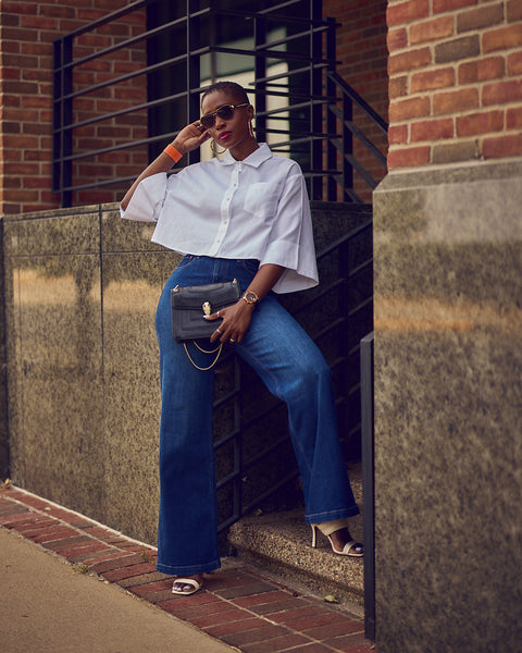 A fashion and style editorial photo showing influencer Farotelle wearing blue wide-leg jeans with a cropped white shirt and white strappy sandals.