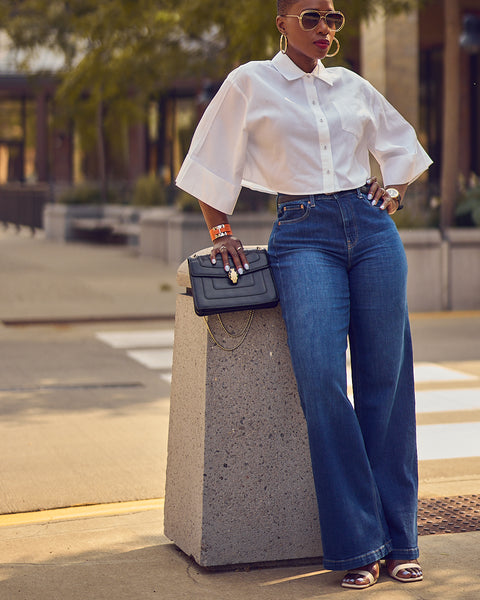 A fashion and style editorial photo showing influencer Farotelle wearing blue wide-leg jeans with a cropped white shirt and white strappy sandals.