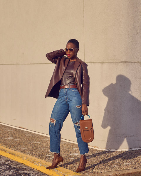 Style blogger Farotelle wearing blue jeans with a brown faux leather blazer. She has on brown pumps and is holding a brown leather handbag.