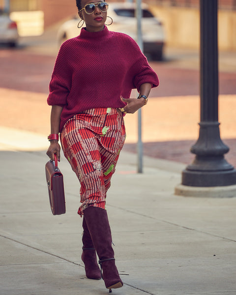 A fashion and style editorial photo showing influencer Farotelle wearing a red printed slip dress with a burgundy sweater and burgundy boots. She is holding a burgundy oblong-shaped leather handbag.