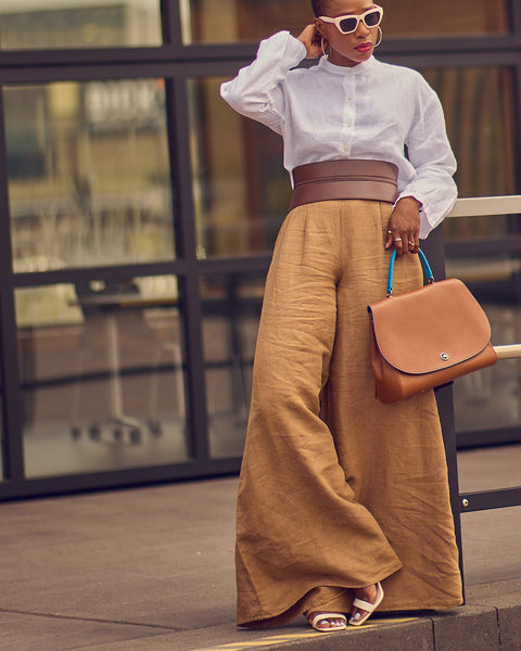 Style influencer Farotelle posing in a neutral outfit consisting of brown wide-leg pants, a white long-sleeve shirt, a wide brown belt, and a brown bag.