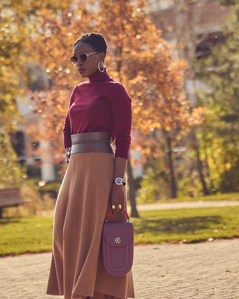 A fashion and style editorial photo showing influencer Farotelle wearing a color-blocked knit outfit. The outfit consists of a camel-colored knit midi skirt, a burgundy turtleneck sweater, burgundy pumps, and a burgundy leather handbag. The look is cinched with a wide brown waist belt.