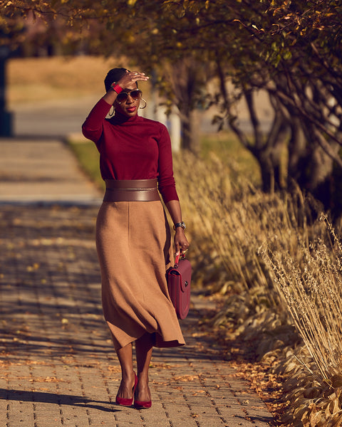 A fashion and style editorial photo showing influencer Farotelle wearing a color-blocked knit outfit. The outfit consists of a camel-colored knit midi skirt, a burgundy turtleneck sweater, burgundy pumps, and a burgundy leather handbag. The look is cinched with a wide brown waist belt.