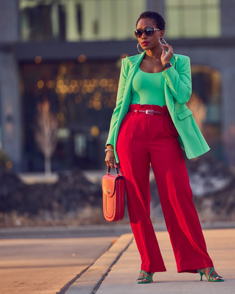 Style influencer Farotelle wearing a red and green color-blocked outfit. She has on high-waisted red pants with a fitted green top and a matching longline green blazer. She's wearing green strappy sandals and is holding a red handbag. She has short natural black and sunglasses.