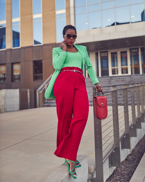 Style influencer Farotelle standing by metal guardrails and wearing a red and green color-blocked outfit. She has on high-waisted red pants with a fitted green top and a matching longline green blazer. She's wearing green strappy sandals and is holding a red handbag. She has short natural black and sunglasses.