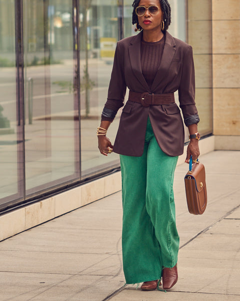 Fashion influencer Farotelle wearing green wide leg jeans with a brown belted blazer. There's a building with windows in the background.