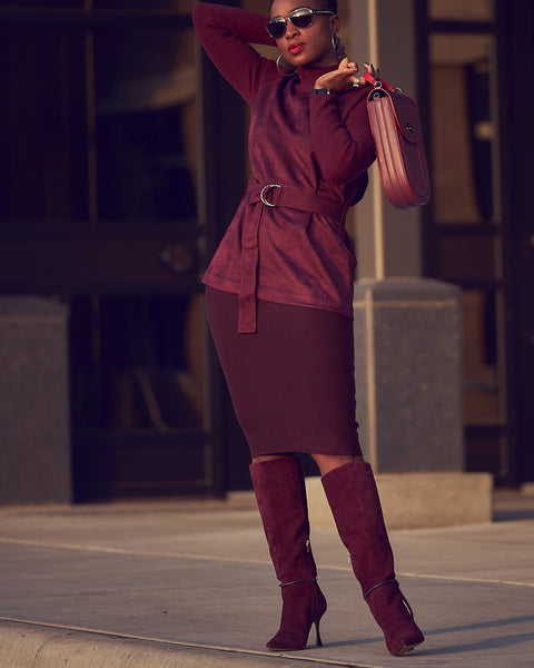 A fashion and style editorial photo showing influencer Farotelle wearing a monochromatic burgundy outfit. The outfit consists of a burgundy knit pencil sweater skirt, a burgundy belted sweater, burgundy knee-high boots, and an oblong-shaped burgundy leather handbag.