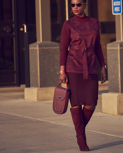 A fashion and style editorial photo showing influencer Farotelle wearing a monochromatic burgundy outfit. The outfit consists of a burgundy knit pencil sweater skirt, a burgundy belted sweater, burgundy knee-high boots, and an oblong-shaped burgundy leather handbag.