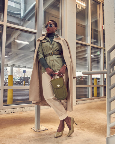 A fashion editorial photo of a woman wearing off-white khaki pants with a belted green jacket. Her shoes are Sam Edelman green heeled pumps and she is holding a green Tomoli handbag. She is standing and shoulder-robing an off-white coat. This is a neutral outfit idea for Fall and Winter style.