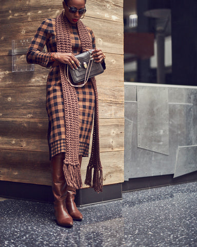 A fashion editorial photo of a woman wearing a brown checkered sweater dress with brown boots and a brown scarf. She is standing by a wall. This is a neutral outfit idea for Fall and Winter style.