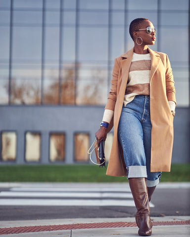 A fashion editorial photo showing a woman wearing cropped skinny jeans with brown boots and an off-white/camel striped sweater. She has on a camel coat and appears to be crossing a street. This is a neutral outfit idea for Fall and Winter style.