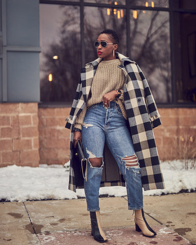 A fashion editorial photo of a woman wearing distressed blue jeans with a beige sweater and a checkered black and beige coat. She has on Alohas bicolor boots and is shoulder-robing her coat. This is a neutral outfit idea for Fall and Winter style.