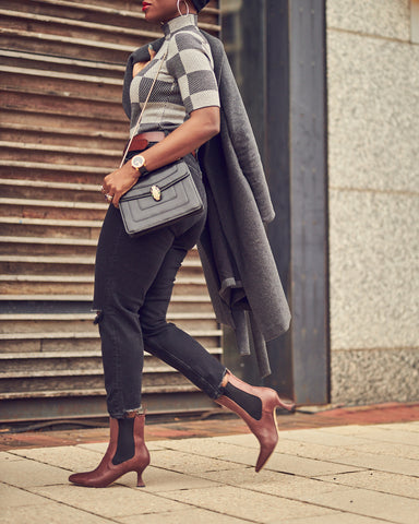 A fashion editorial photo of a woman wearing slim black jeans with a grey checkered sweater and holding a grey coat. She has on brown ankle boots and appears to be walking. This is a neutral outfit idea for Fall and Winter.