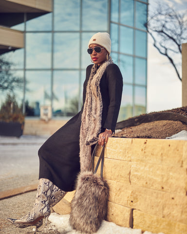 A fashion editorial photo showing a woman wearing a black dress with snakeprint boots and a grey/beige fur scarf. She is holding a grey fur bag and has on a winter hat. This is a neutral outfit idea for Fall and Winter style.
