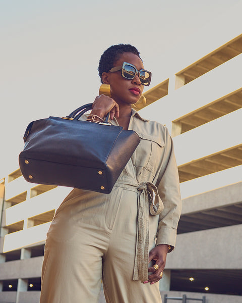 Farotelle wearing neutral utility jumpsuit, sunglasses, large gold earrings and holding black leather tote bag as Business Casual Spring Summer Workwear Outfit.
