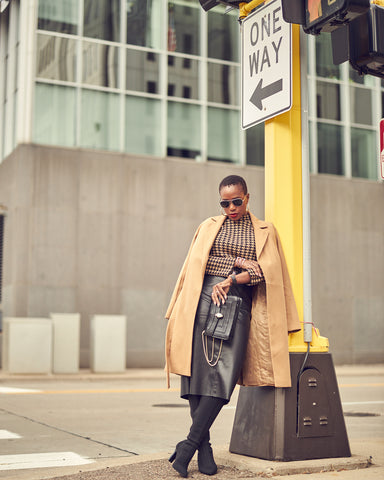 A fashion editorial photo of woman wearing a black leather skirt with tall black boots and a beige houndstooth sweater. Her coat hangs over her shoulders and she appears to be by a street sign post. This is a neutral outfit idea for Fall and Winter.