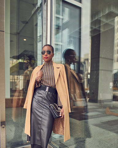 A fashion editorial photo of woman wearing a black leather skirt with a beige houndstooth sweater. Her coat hangs over her shoulders and she is standing by glass windows. This is a neutral outfit idea for Fall and Winter.