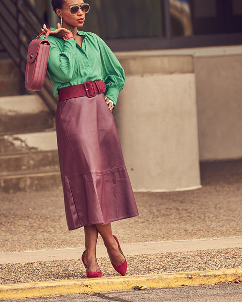 A black woman in a standing position wearing a burgundy faux leather skirt with a contrasting green blouse. She's also wearing burgundy heeled pumps and has a burgundy handbag.