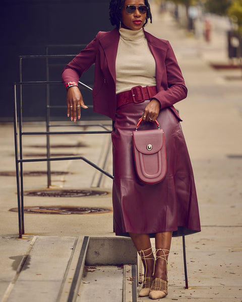 A black woman in a standing position wearing a matching burgundy skirt and cropped jacket. She's holding a small burgundy handbag and wearing off-white heeled lace-up shoes. This is a monochrome office look.