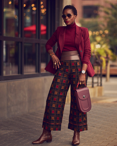A black woman in a standing position and wearing printed pants with a solid burgundy turtleneck and blazer. Her shoes and belt are made of brown leather. This is a Fall office look.