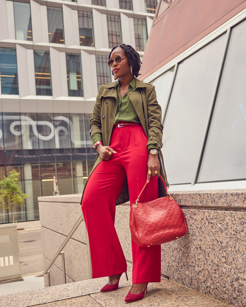 A black woman wearing red wide leg pants with an olive shirt and matching olive trench coat. She's wearing pump heels and holding a red Louis Vuitton handbag.