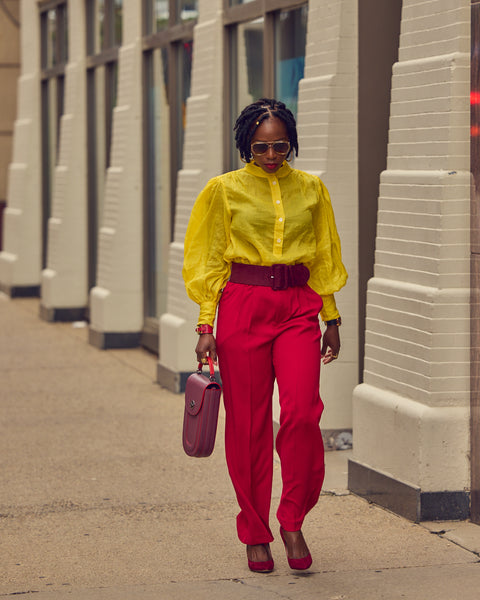 A black woman walking outside and wearing a bright color-blocked outfit. The outfit consists of wide leg red pants, a yellow blouse, and matching burgundy heels and belt. Her hair is styled in locs.