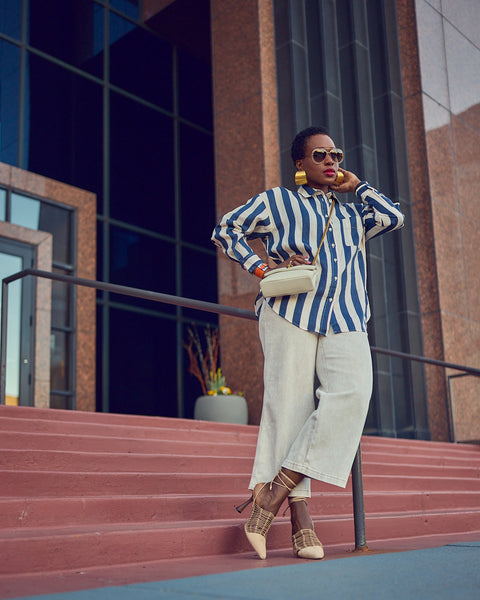 Style influencer Farotelle wearing an elevated casual Spring outfit. The outfit consists of an oversized striped button-down shirt with light denim culottes. She is wearing a chain crossbody bag and lace-up heels. She is standing on stairs.