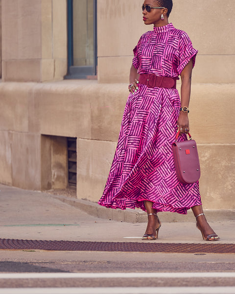 Style influencer Farotelle wearing a matching pink skirt and blouse ensemble in a silky, print material. She has on silver strappy sandals and is holding a burgundy handbag that matches the pattern of the fabric. The look is cinched with a wide burgundy belt.