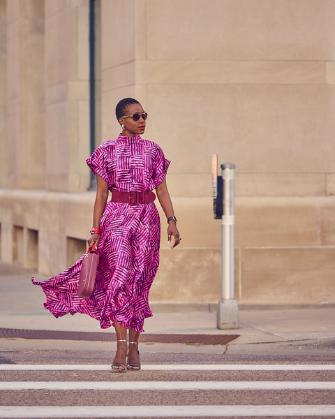 Style influencer Farotelle wearing a matching pink skirt and blouse ensemble in a silky, print material. She has on silver strappy sandals and is holding a burgundy handbag that matches the pattern of the fabric. The look is cinched with a wide burgundy belt. She is standing at a street light.